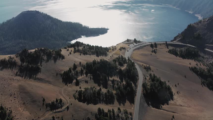 A winding road climbs a mountainside passing through a landscape of dry grasses and scattered trees offering views of a large lake and distant mountains