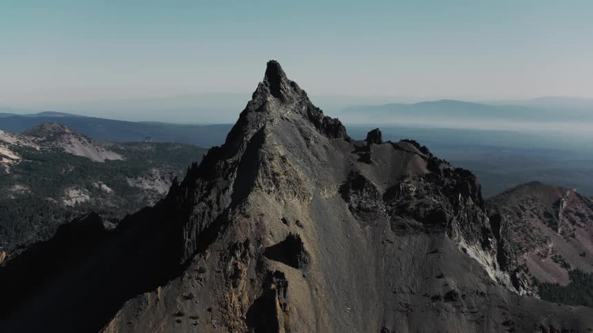 A sharp rocky peak rises dramatically above a vast landscape of rolling hills and distant mountains its dark surface contrasting sharply with the bright blue sky