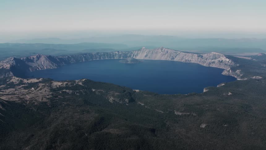 An aerial view of crater lake a deep blue caldera lake surrounded by steep cliffs and forested terrain with wizard island visible in the center