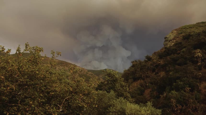 A plume of smoke rises from a wildfire burning in a mountainous region with dark clouds gathering overhead