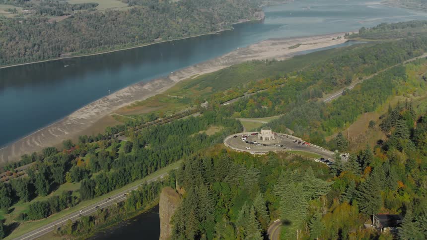 Vehicles drive toward and from a building on a cliff overlooking a lake in forested mountainous terrain on a foggy day