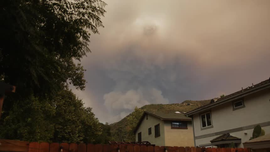 A large plume of smoke billows from a hillside fire casting a hazy shadow over a residential neighborhood with houses and trees