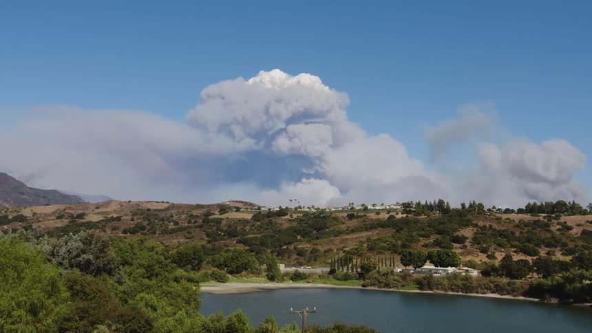 Rural landscape with green hills and a calm lake in the foreground and white smoke rising on the horizon