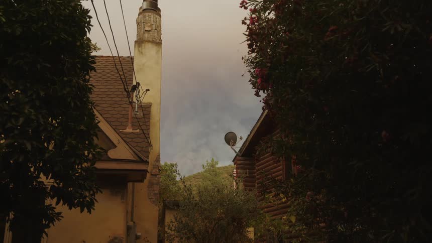 A hazy daytime shot between two houses one with a chimney looking towards a distant treeline and a that sky is overcast and theres a satellite dish on the house to the right