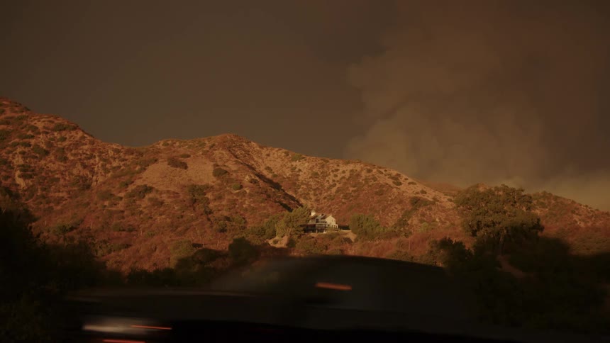 House on the side of a mountain as traffic passes and smoke rises in the distance