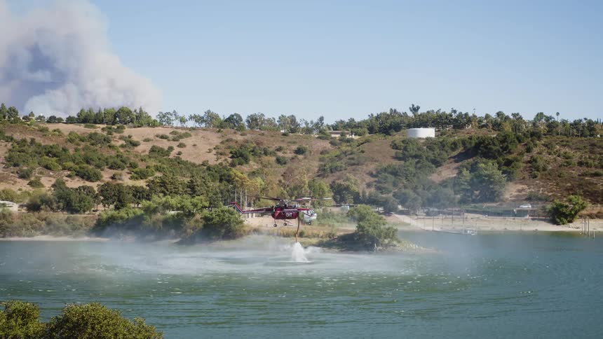 Firefighting helicopter fills up with water from a lake on the opposite side of a hill from the source of a large smoke cloud