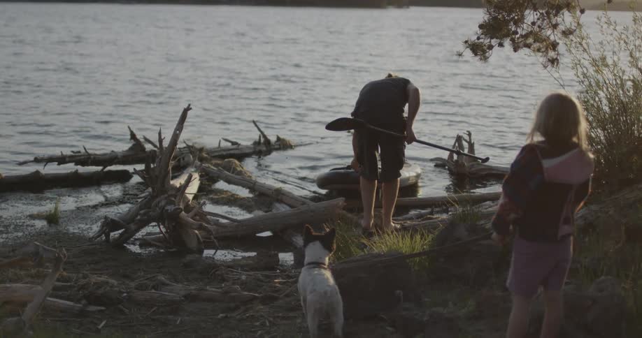 Man putting kayak in water with a child and dog watching him