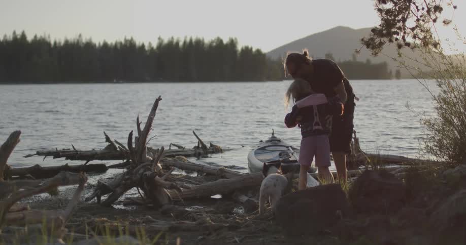 A man is lifting a young child as they stand near the edge of a lake