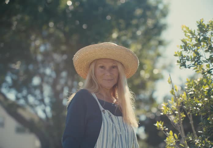 A smiling older woman with long blonde hair wearing overalls and a straw hat looks directly at the camera in a garden setting