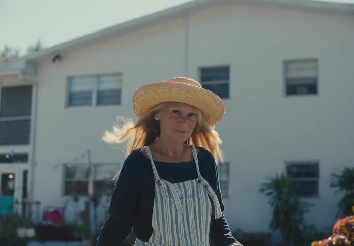 A lady wearing a straw hat and striped overalls stands in front of a white house with plants visible in the background