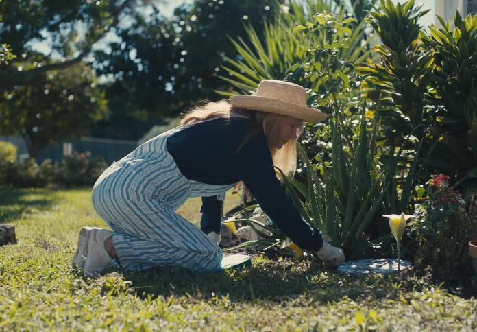Woman wearing a hat and gloves crouching on the grass and picking dry leaves from plants