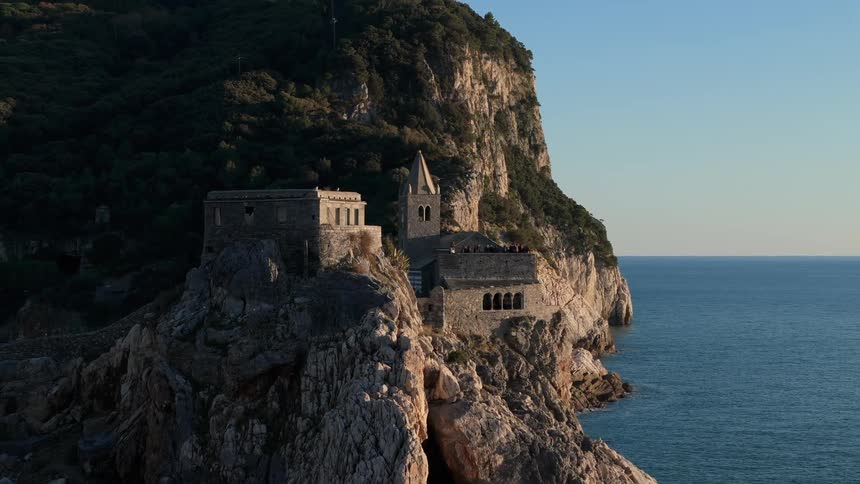 A historic stone building possibly a church or fortress is perched dramatically on a rocky cliff overlooking a calm expansive sea with a clear sky in the background