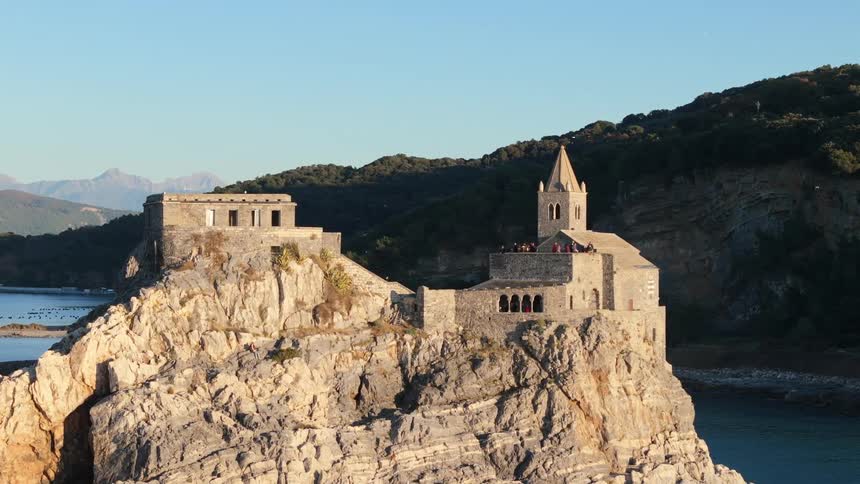 A historic church and buildings stand perched on a rocky cliff overlooking the calm blue waters of the mediterranean sea at sunset