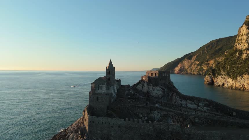 A historic church and buildings stand perched on a rocky cliff overlooking the calm blue waters of the mediterranean sea at sunset