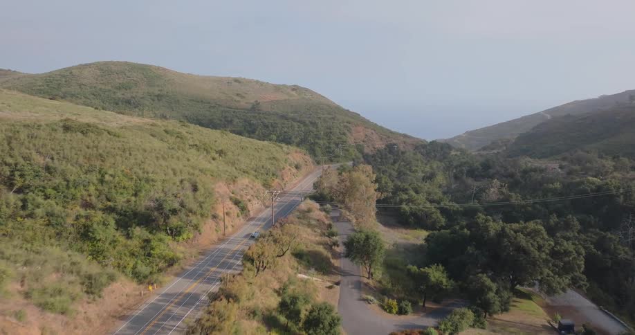 A two lane highway winds through a hilly landscape with patches of trees and shrubs lining the roadside and a single car driving along it