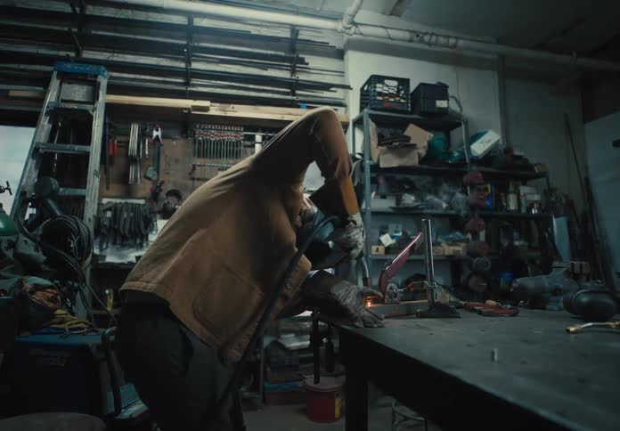 A person is welding a metal piece on a workbench in a cluttered workshop filled with various tools and equipment