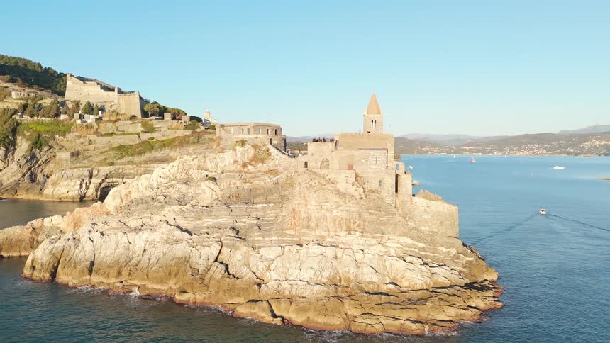A bird flies over the rocky outcrop of a town overlooking the blue sea with distant hills