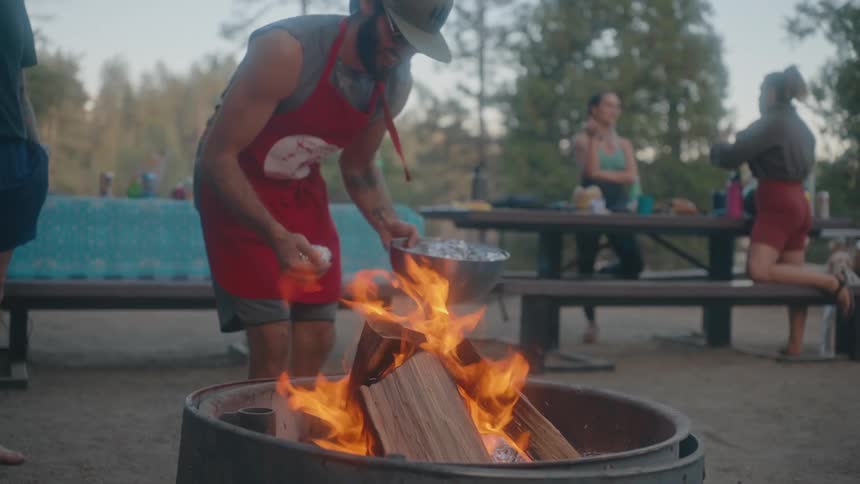 A man wearing a red apron cooks food in foil packets over a bonfire while others socialize nearby at a campsite