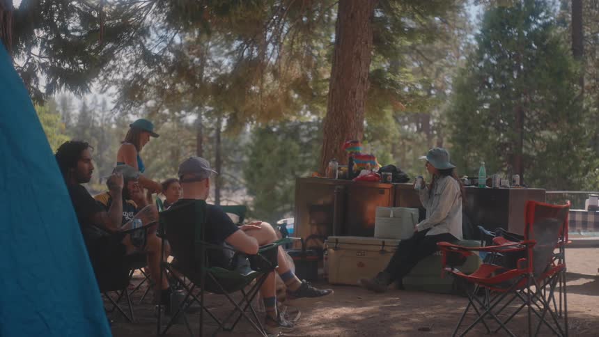 A group of people is gathered around a campsite with a tent camping chairs a dog and various camping supplies under tall trees in a forested area