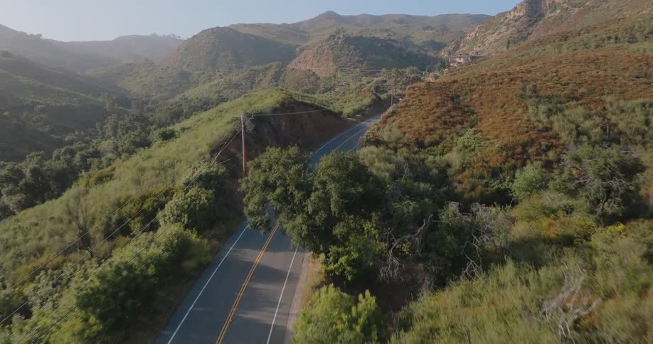 A silver car drives along a two lane road that winds through rolling hills covered in green vegetation with power lines running alongside