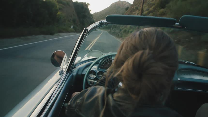 Woman driving a classic car on a road surrounded by mountains