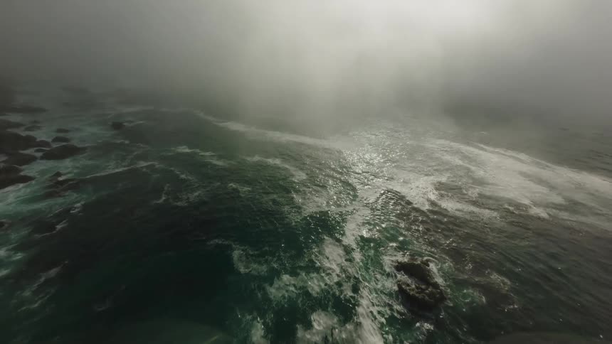 A rugged cliff face drops steeply into a turbulent sea with crashing waves and a thick layer of fog obscuring the view beyond