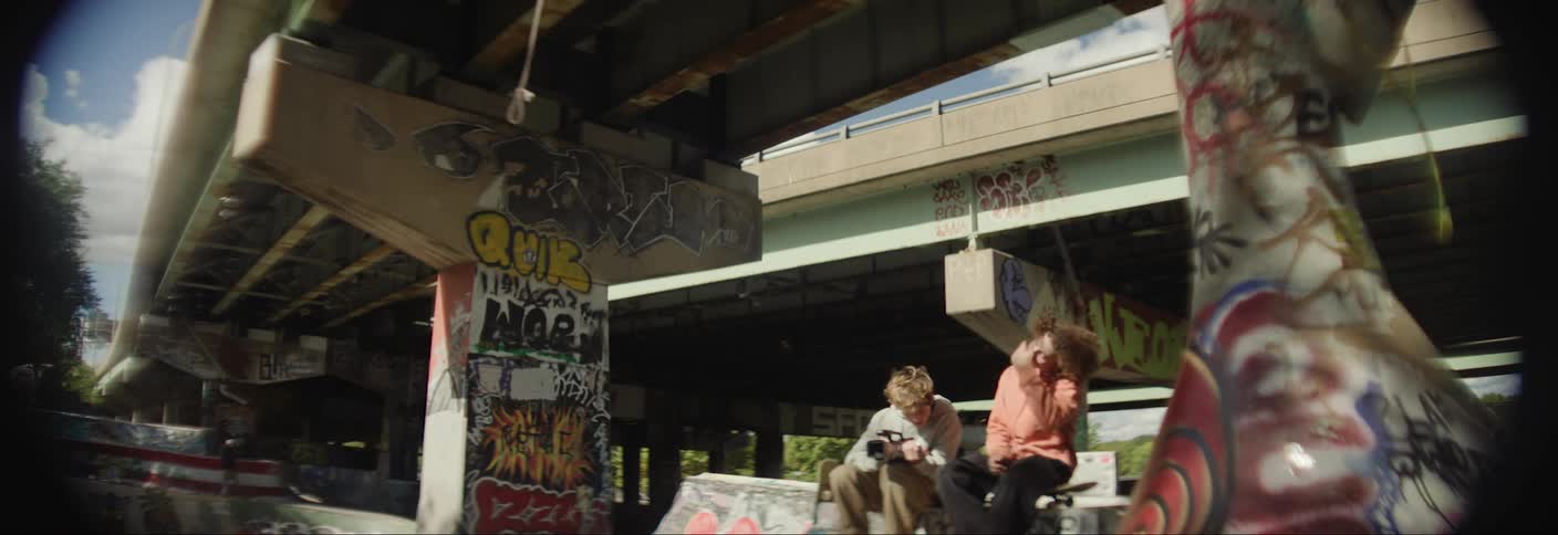 Two friends sitting on a bench under an overpass and watching a professional camera