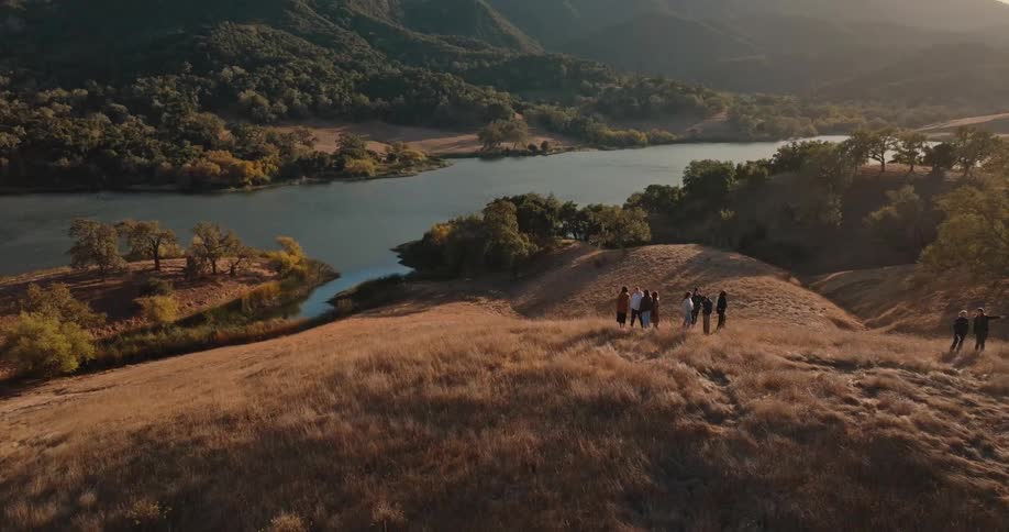 A group of people is walking on a grassy hill overlooking a winding river and a forested landscape