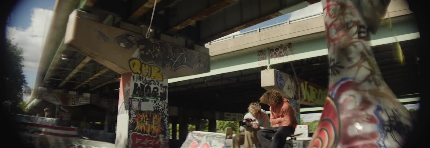Two teenagers sitting on a bench below an overpass talking