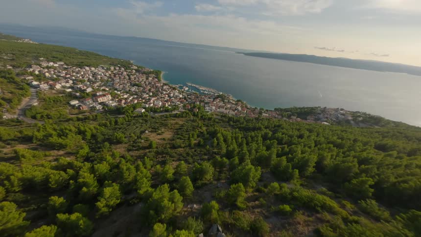 A coastal town with white buildings and a harbor is nestled among green hills and forests with a clear blue sea stretching out beyond