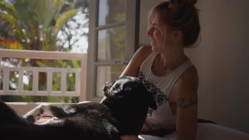 Woman smiles as her dog gets up from lying with her on a cushion near open balcony doors on a summer day