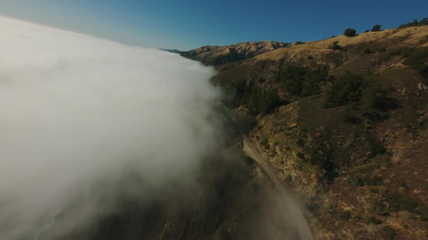 A winding road follows a coastal cliffside with a thick layer of fog rolling in from the ocean below and a clear blue sky visible above
