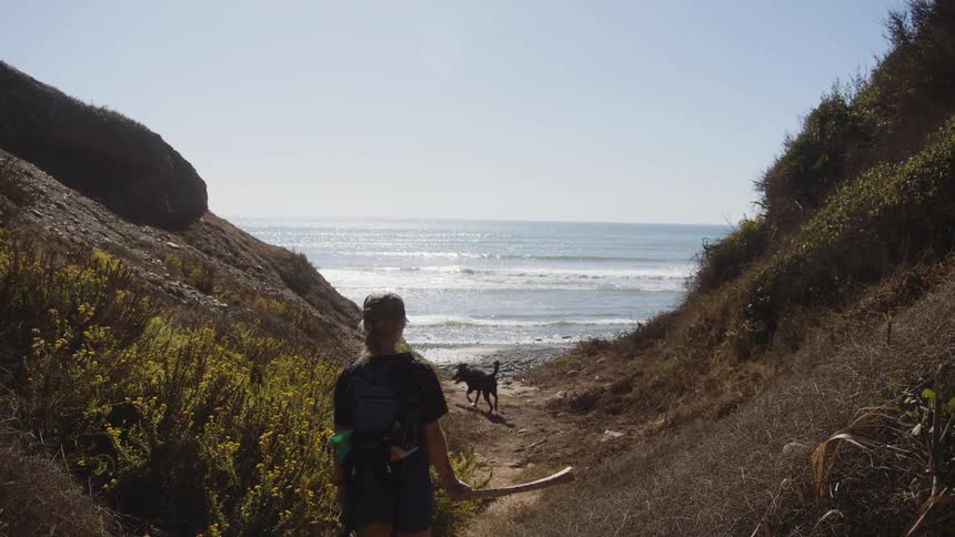 A woman wearing a cap a black shirt shorts and a backpack walks along a narrow dirt path bordered by yellow wildflowers and rocky slopes heading toward a tranquil ocean horizon with a dog following closely behind
