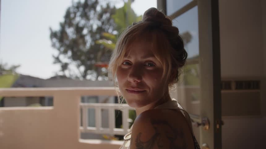 A woman with a bun hairstyle and a tattoo on the shoulder stands by an open door looking out at a sunlit outdoor