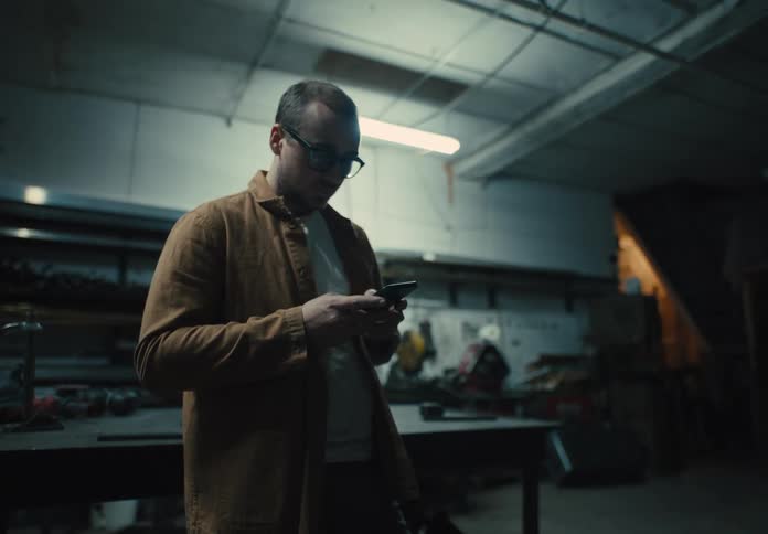 A man in a workshop surrounded by tools is texting on his phone