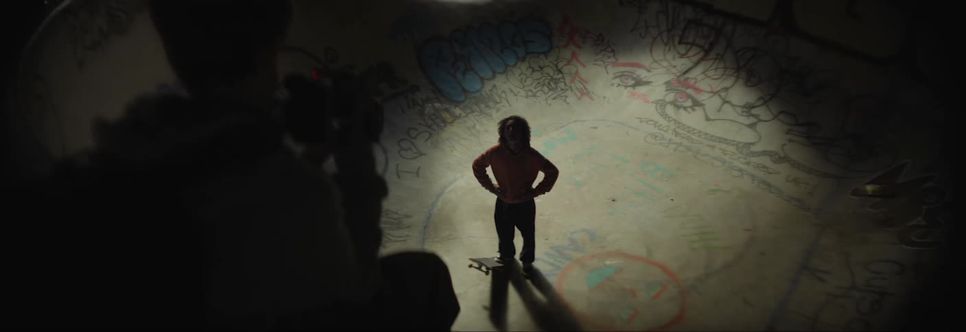 A young teen stands in a graffiticovered skate bowl with a skateboard illuminated by a spotlight while being filmed by someone in the foreground
