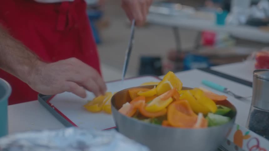 A chef wearing a red apron uses a knife to slice yellow bell peppers on a cutting board with a bowl of chopped vegetables nearby