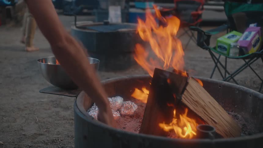 Campfire with intense flames while a person places food wrapped in aluminum foil on the coals