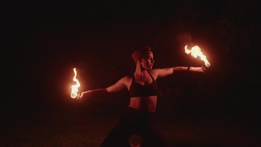 A woman is performing a fire dance at night holding two flaming torches while kneeling on the ground