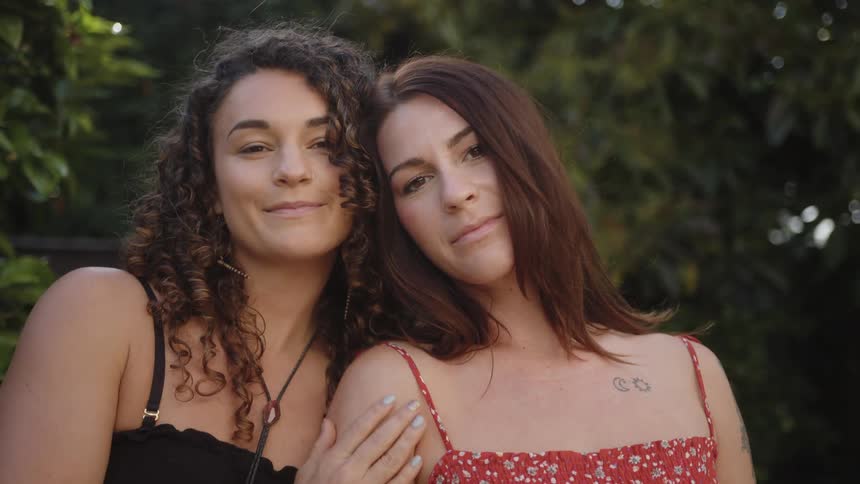 Two women outdoors one with curly hair wearing a black top and the other with straight hair wearing a red floral dress both smiling softly with a relaxed demeanor