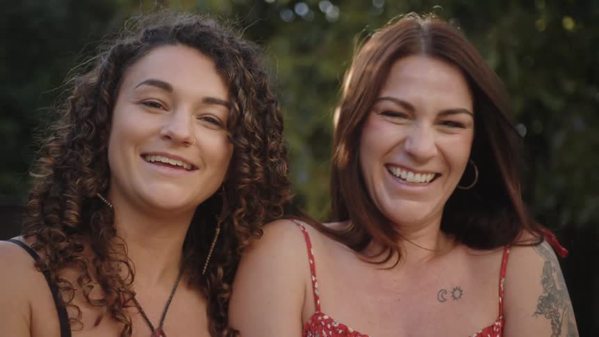 Two young women with curly and straight hair respectively sit close together outdoors laughing heartily and sharing a lighthearted moment