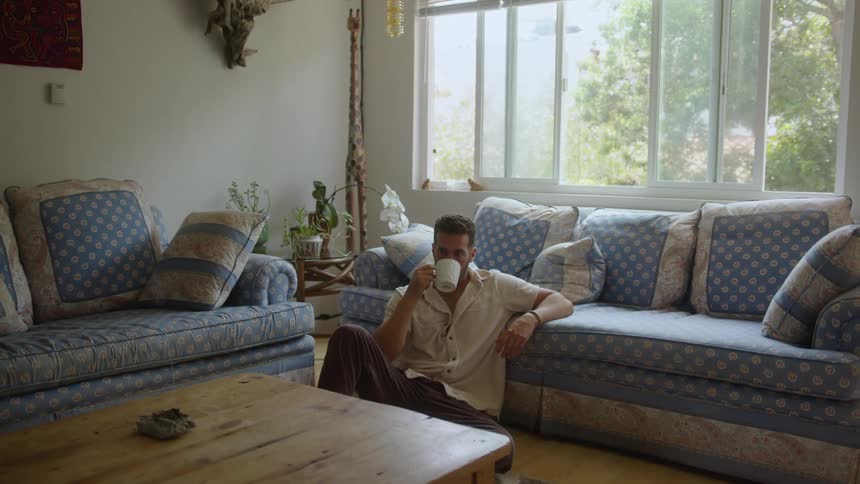 A man sits on the floor of a bright cozy living room leaning against a bluepatterned sofa sipping from a white mug with sunlight streaming through large windows