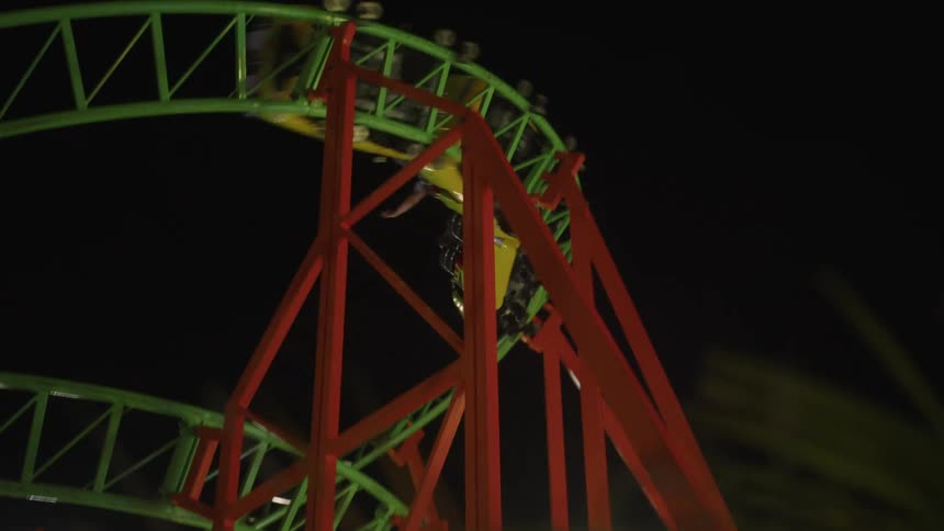 A brightly lit roller coaster at night features green and orange tracks and a yellow train carrying passengers with their hands raised in excitement