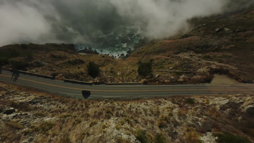 A winding road cuts through a rugged mountainous landscape with sparse vegetation leading towards a misty coastline where waves crash against the rocks below