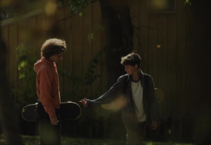 Two teenagers with skateboards are talking near a wooden fence