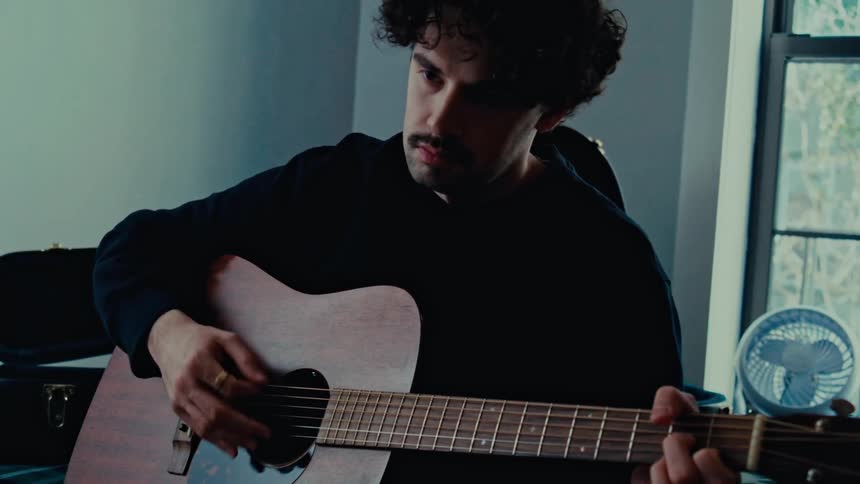 Male guitarist sitting near a window plays an acoustic guitar