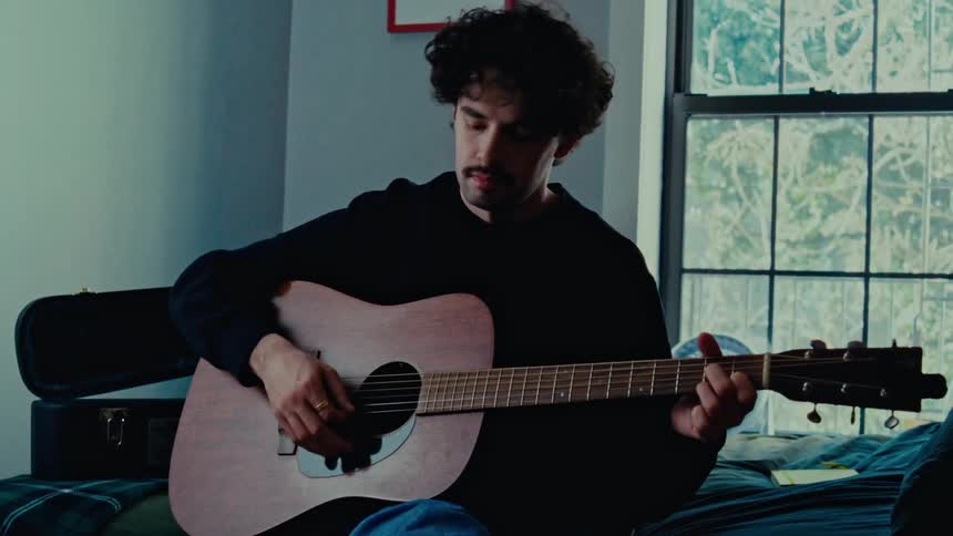 A man with curly hair a mustache and a black sweater sits on the bed strumming an acoustic guitar in the room