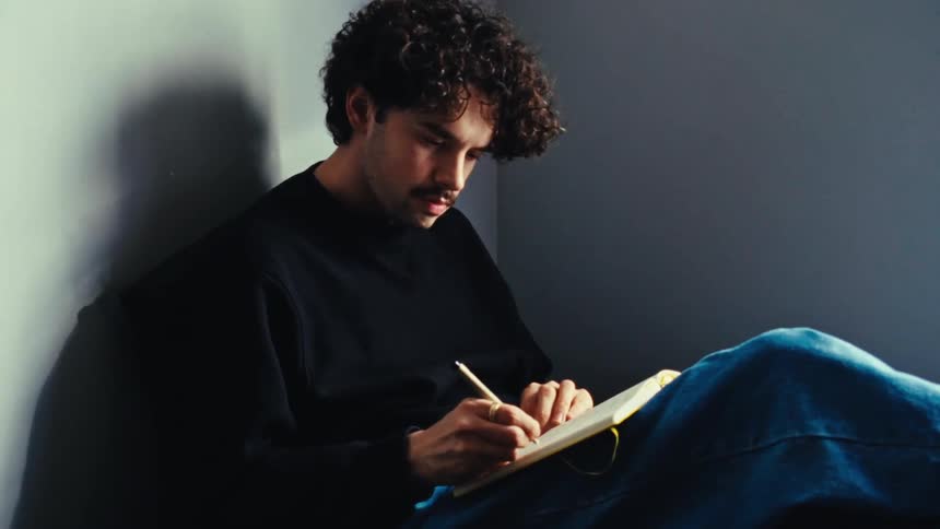A man with curly hair wearing a black sweater and jeans writes down notes in a notebook in the corner of a room with gray walls