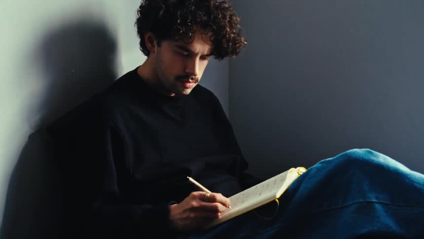 A man with a mustache wearing a black sweater and jeans writes down notes in a notebook in the corner of a room with gray walls
