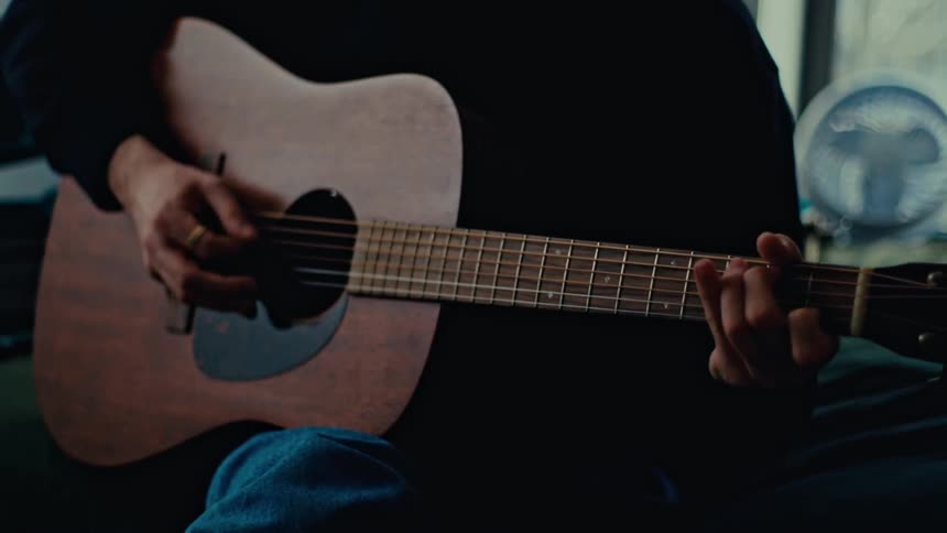 A persons hands are shown playing a brown acoustic guitar while wearing a black shirt and blue jeans